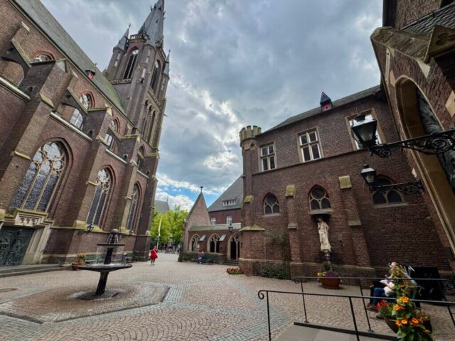Innenhof der Marienbasilika Kevelaer mit Blick auf die neugotische Fassade und den hohen Kirchturm. Umgeben von Backsteingebäuden mit Spitzbogenfenstern und kleinen Türmen. In der Mitte des gepflasterten Platzes steht ein Brunnen, rechts eine Marienstatue in einer Nische. Blumen schmücken den Vorplatz, eine Frau sitzt auf einer Bank. Der Himmel ist wolkenverhangen.