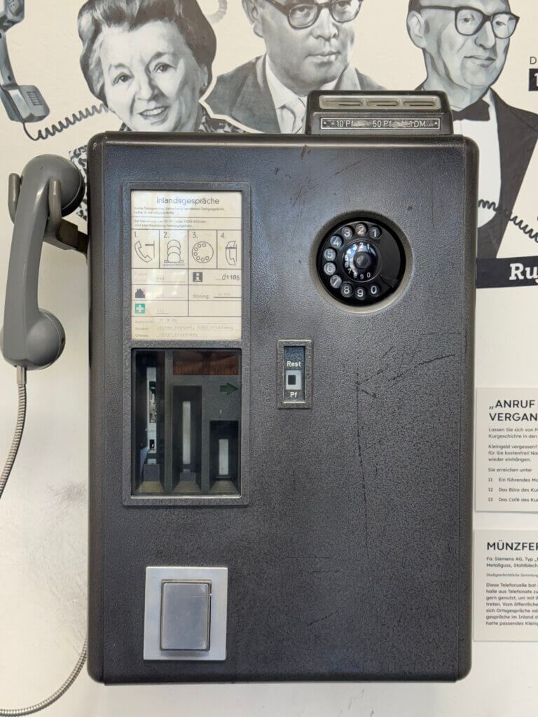 Historisches Münztelefon der Deutschen Bundespost mit Wählscheibe und grauem Hörer, ausgestellt vor einer Wand mit Schwarz-Weiß-Porträts bekannter Persönlichkeiten; daneben eine Infotafel mit der Überschrift ‚Münzfernsprecher‘, fotografiert in einem Museum. Telefonzellen Nostalgie Kommunikationskultur